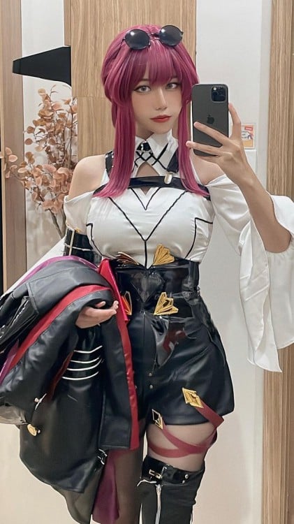 A pretty Korean girl taking a selfie while wearing a steampunk-style costume. She has fucia coloured hair with sunglasses perched on her head. She wears a white top and black leather shorts with a cinched waist to accentuate her already large bosom. She carries what looks like a heavy coat over one arm and appears to be standing in a hotel lobby.