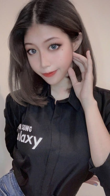 Pretty Korean girl with shoulder length brown hair, wearing a black Samsung button shirt. She smiles at the camera holding her left hand to her cheek.