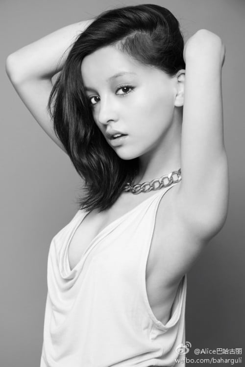 A black and white image of a pretty Asian woman with medium length black hair. She stands sideways to the camera with her hands behind her head holding her hair back. She wears a white sleeveless v-neck top with a chain necklace around her neck.