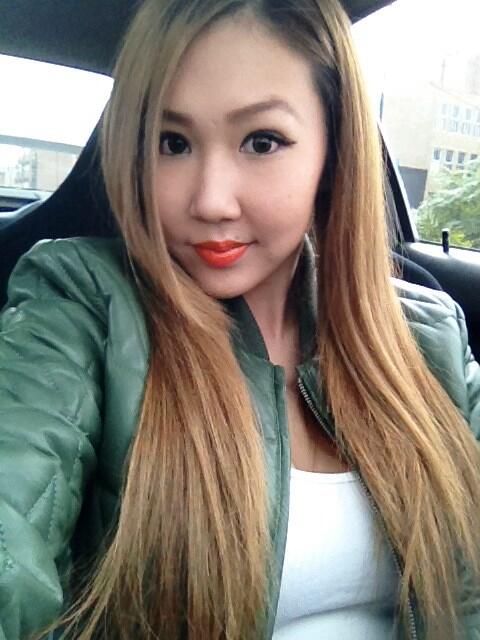 A selfie of a pretty Asian girl with long dark blonde hair, taken inside a car. She smirks at the camera, and wears a green jacket and white, scoop-necked top.